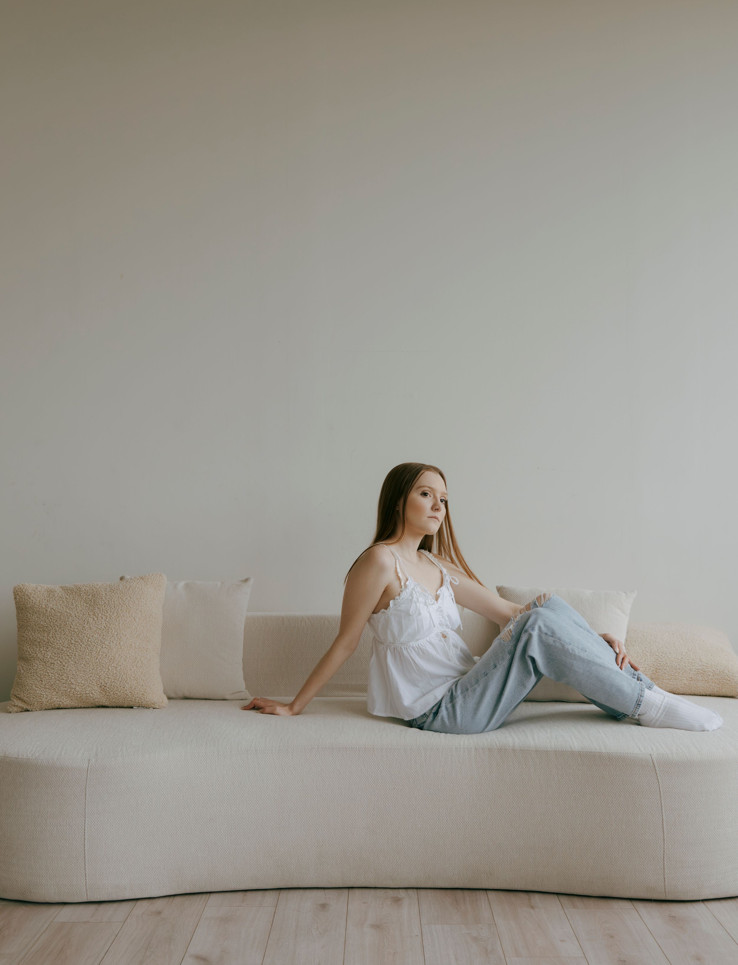 Young woman in white camisole and jeans relaxing on a cream curved sofa with pillows.