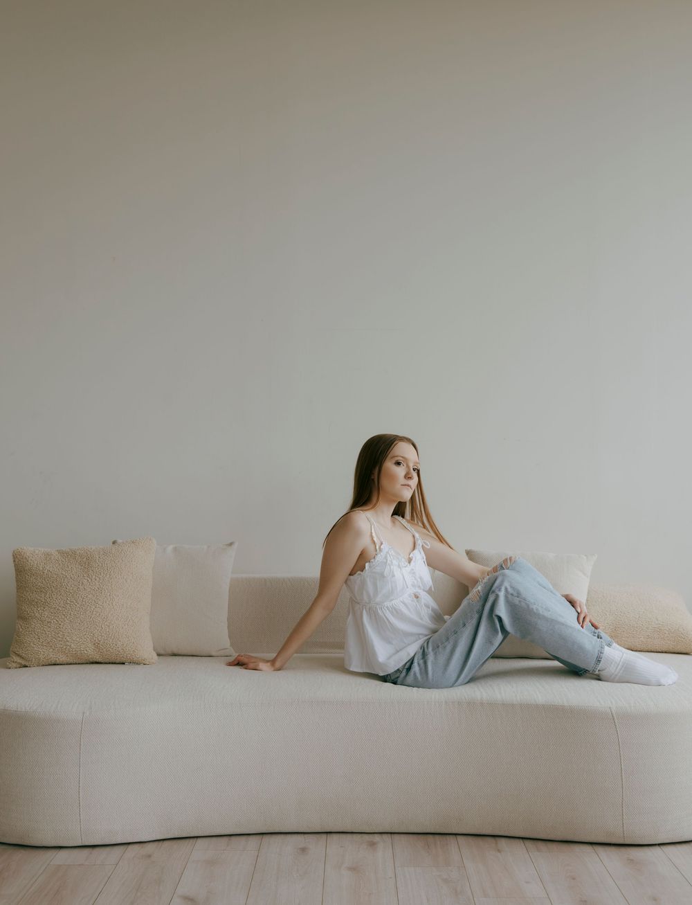 Young woman in white camisole and jeans relaxing on a cream curved sofa with pillows.