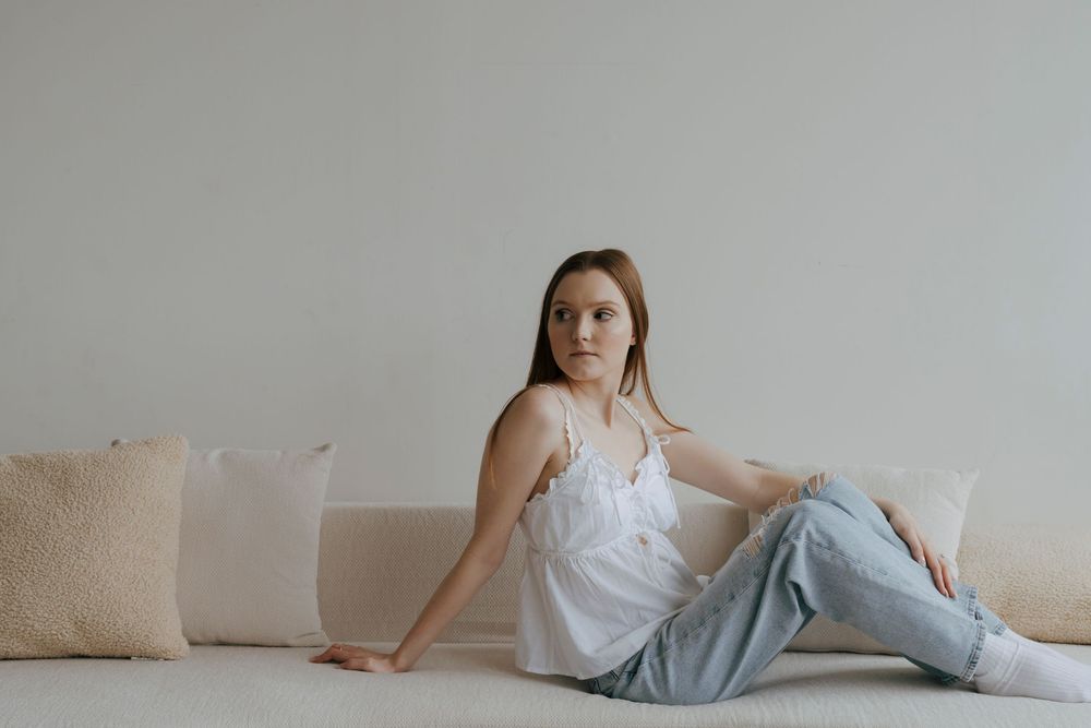 Young woman in white camisole and blue jeans sitting on floor leaning against a beige sofa.