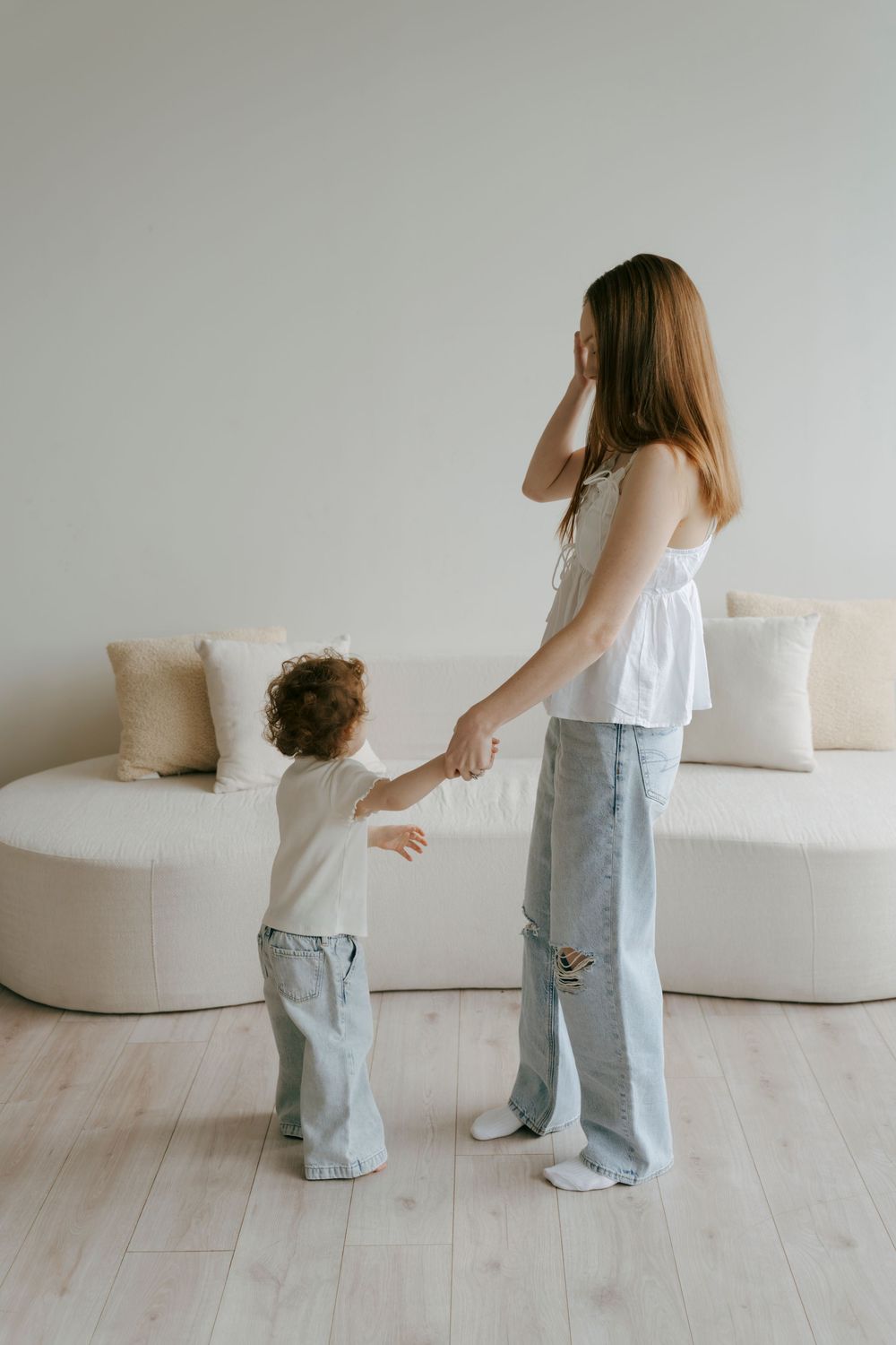 A young mother in jeans holds hands with her toddler in a bright, minimalist white living room.