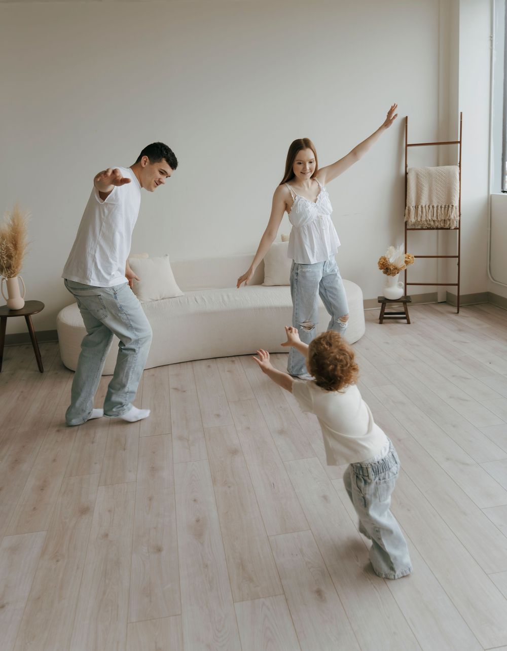 A happy family of three dancing and playing together in a bright modern living room with light wood floors.