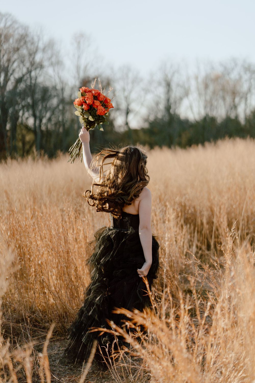 A figure in a dark dress holds up a vibrant orange bouquet while standing in a golden field.