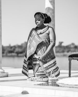 Woman in traditional African print dress standing gracefully by waterfront in black and white photography.