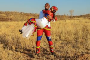 A couple in traditional African attire embraces playfully in a golden field at sunset.