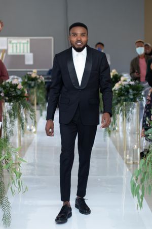 Groom in classic black tuxedo walking down aisle decorated with greenery and white flowers.