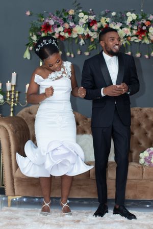 A wedding couple shares a joyful moment posing together against a floral backdrop and brown tufted couch.