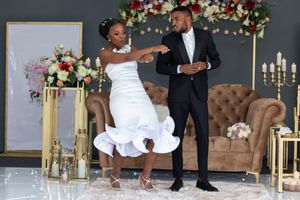 Newlyweds share a playful moment in front of a floral arch and brown couch.