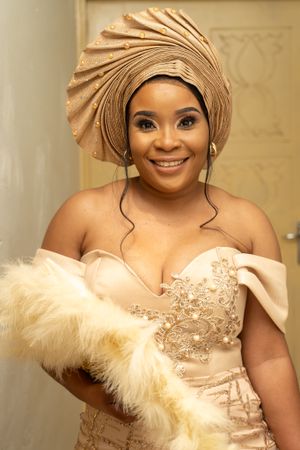 A woman in a champagne-colored dress and matching gele headwrap holds a feathered fan.