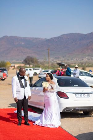 A couple poses on a red carpet with mountains and parked cars in the background at an outdoor wedding.