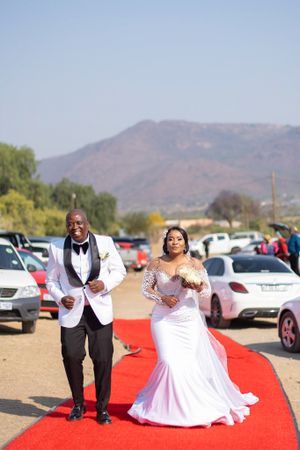 A couple walks down an outdoor red carpet aisle with mountains and parked cars in the scenic background.