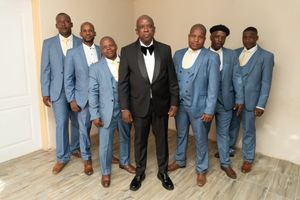Groomsmen in light blue suits pose with groom in black tuxedo against neutral wall.