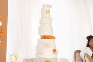Elegant white tiered wedding cake with orange floral accents against white backdrop.