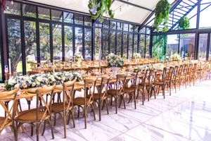 Long rustic wooden tables set for a wedding reception in a greenhouse-style venue with natural lighting.