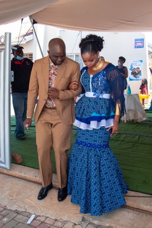 Person in brown suit escorting another wearing blue patterned dress at formal event.