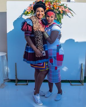 Two friends pose together in colorful traditional African-inspired dresses against a floral backdrop.