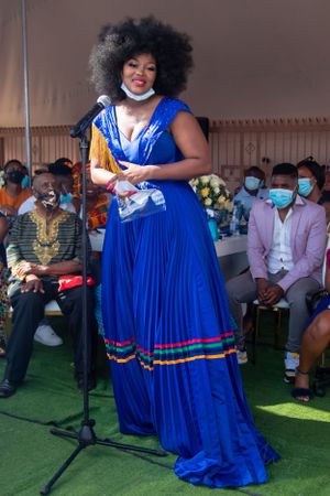 Performer in flowing royal blue pleated gown with rainbow trim speaking at outdoor event.