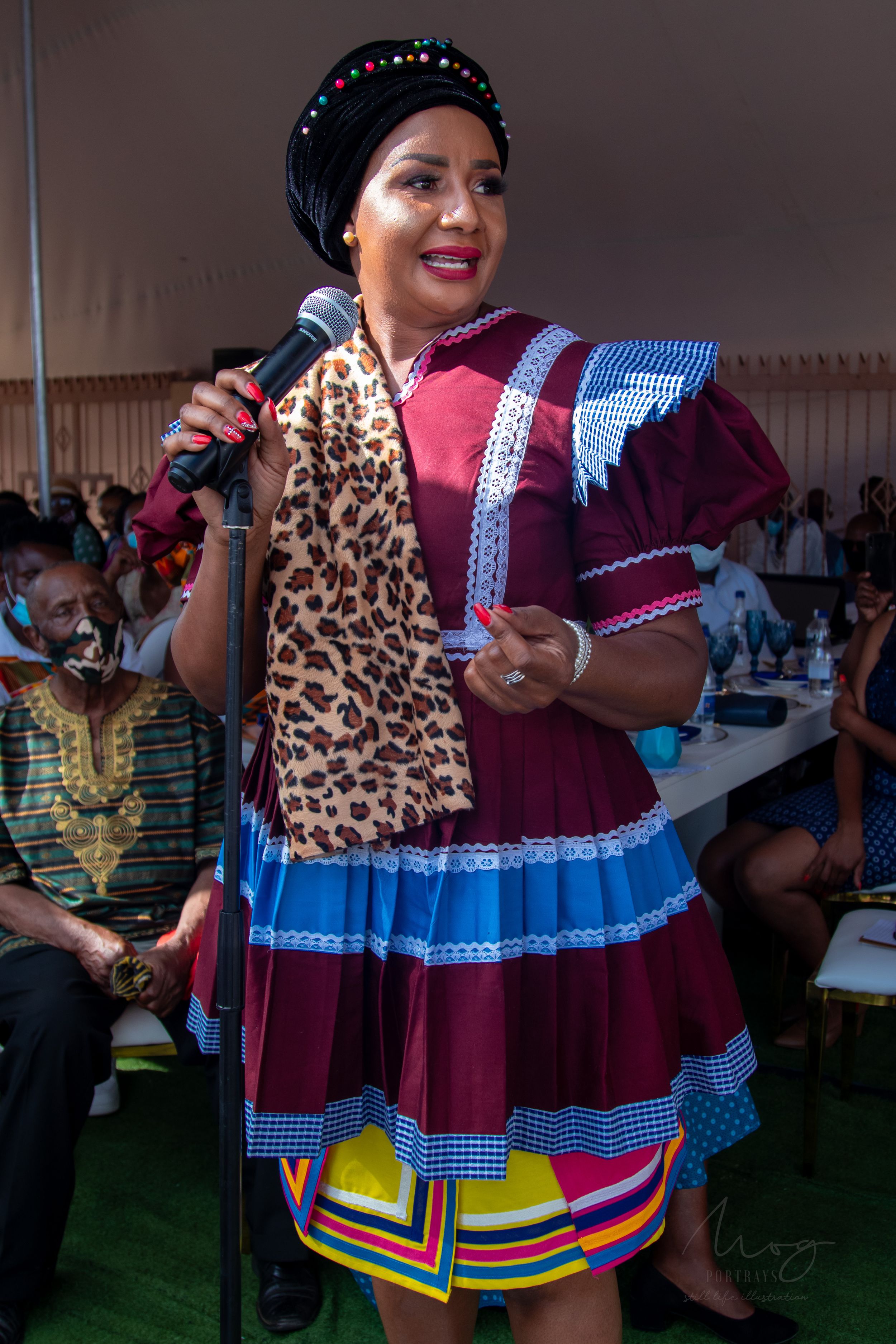 Speaker wearing colorful traditional dress with leopard print scarf at outdoor gathering.