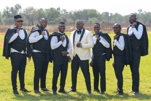 Groomsmen in black suits pose with the groom in white on a sunny wedding day.