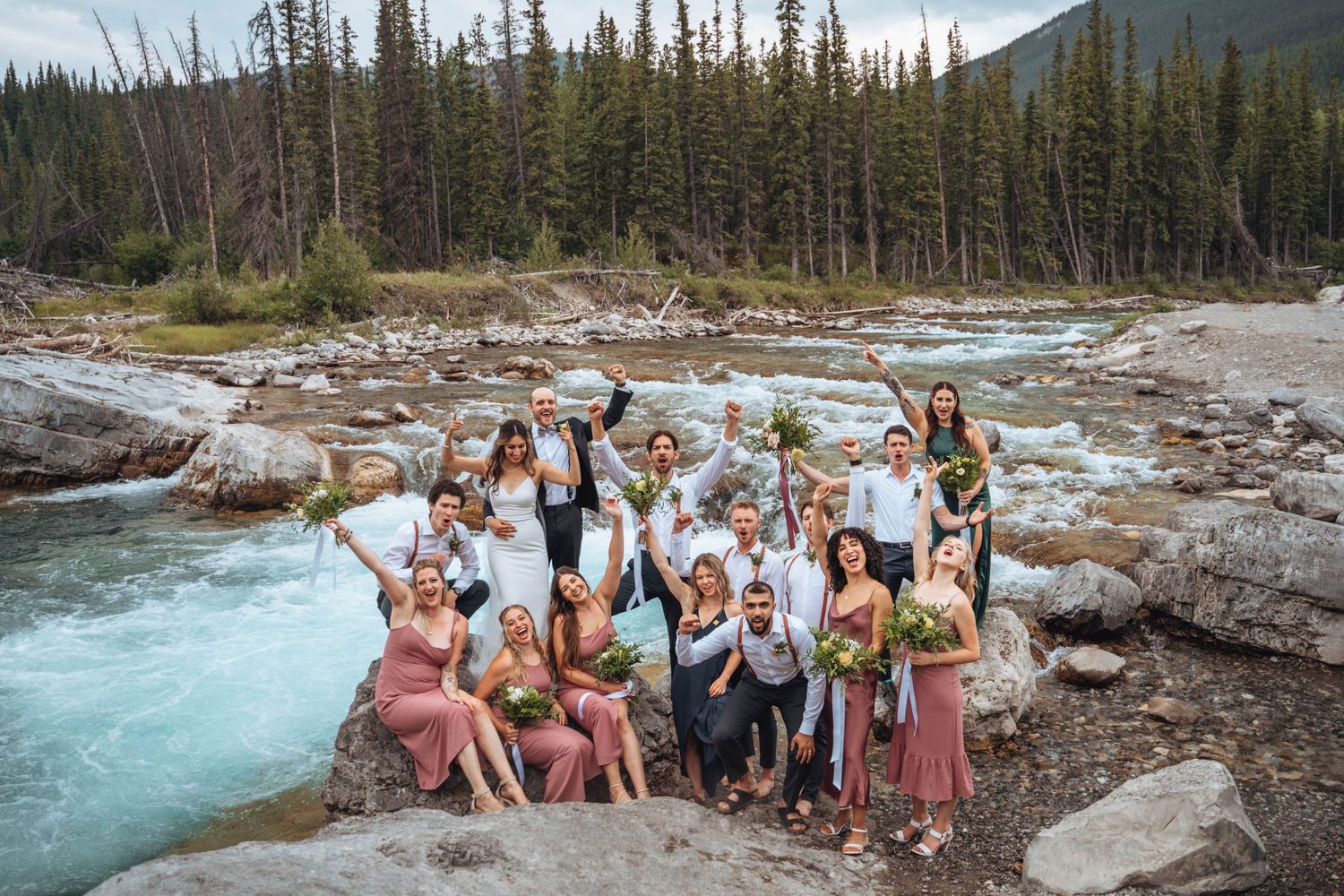 The wedding party poses on rocks by a rushing river, surrounded by forest. Banff wedding.