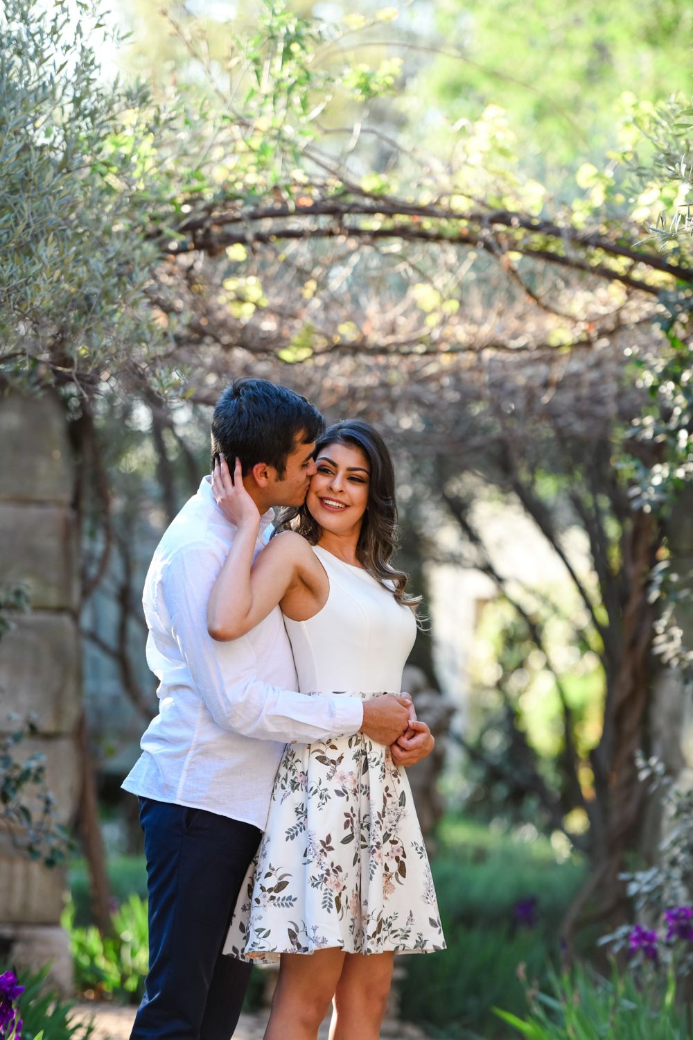 Man kissing his fiancée on the cheek while standing under a garden trellis.