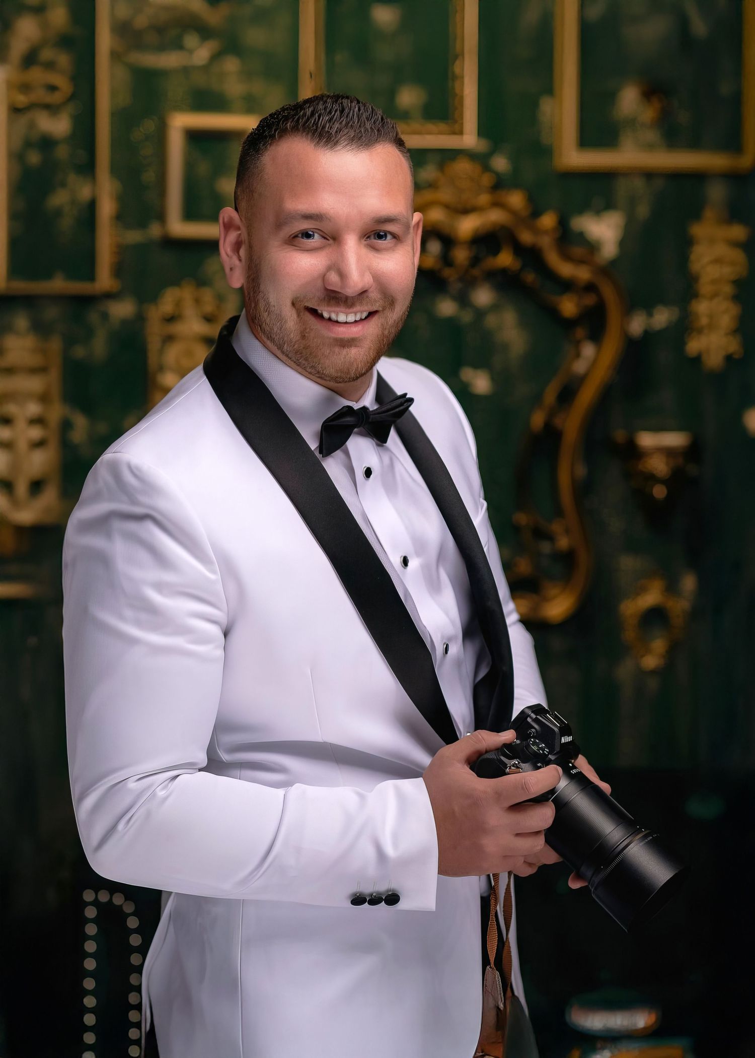 Professional photographer in white tuxedo jacket with black lapels holds camera against ornate golden backdrop.