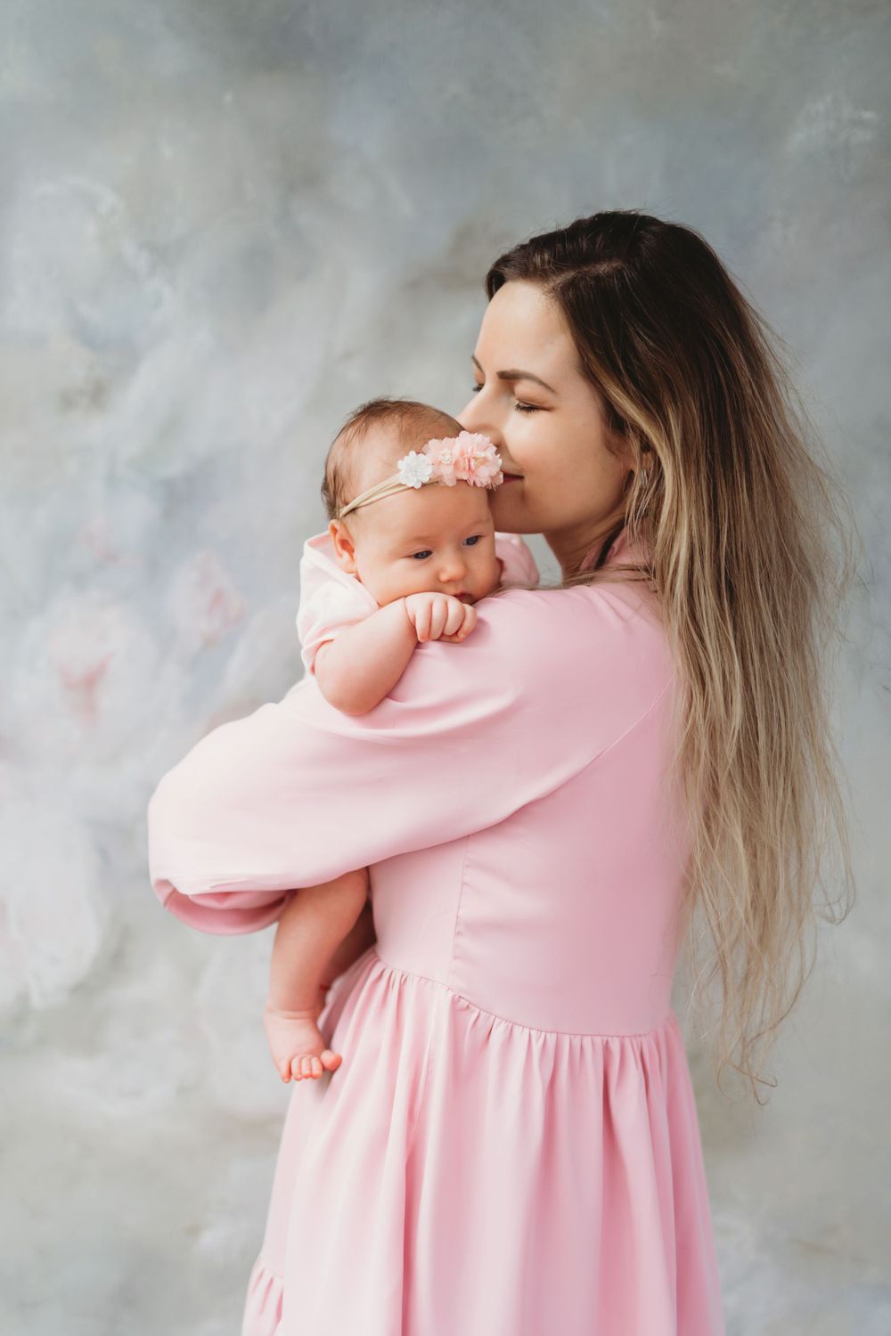 Mother in pink dress cuddles and kisses newborn baby against gray backdrop in studio portrait. Bellevue studio, WA