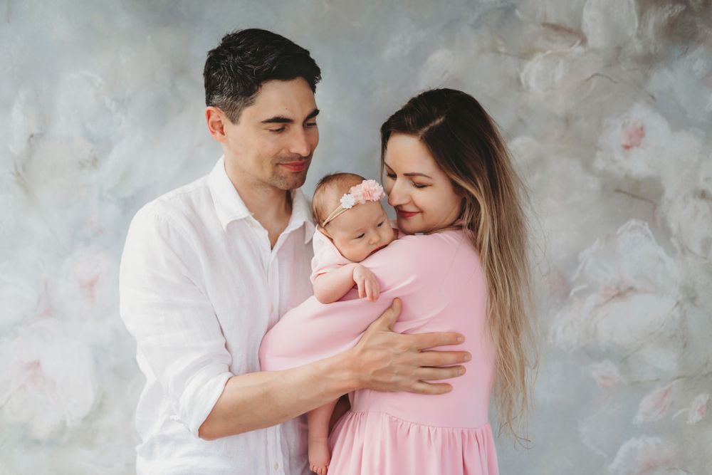 A couple in white and pink outfits embrace their newborn baby against a textured gray background in a studio portrait. Bellevue studio, WA