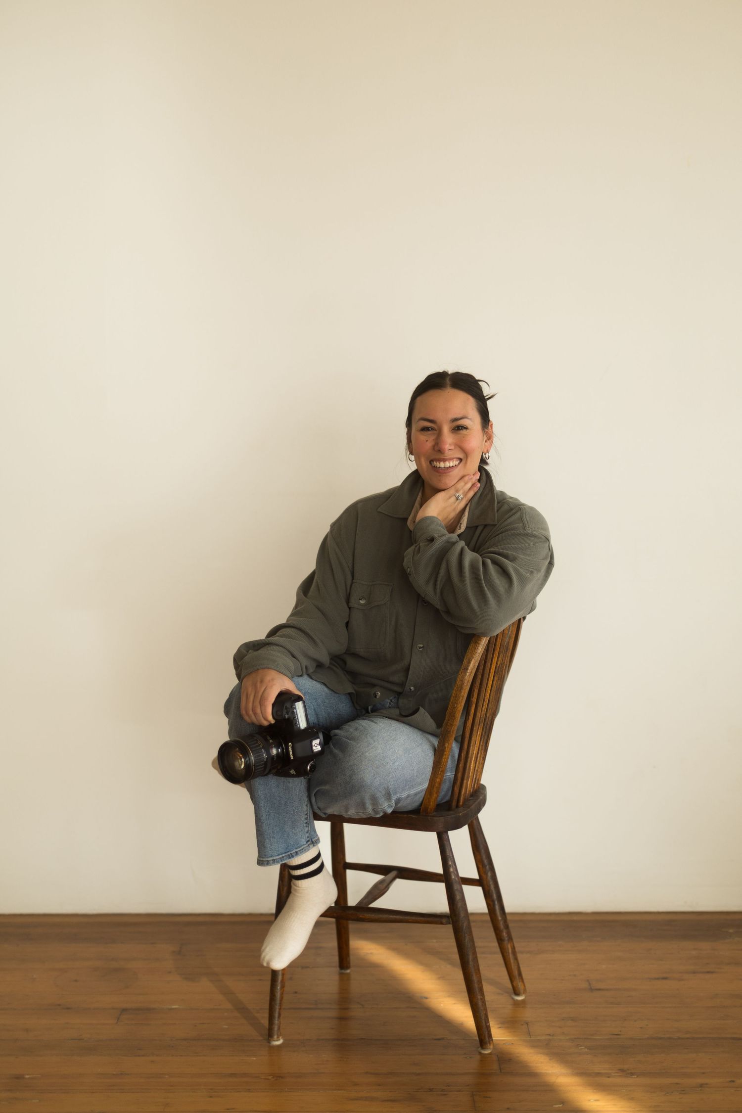 Photographer in casual attire sits on wooden chair holding camera against neutral wall with warm lighting.