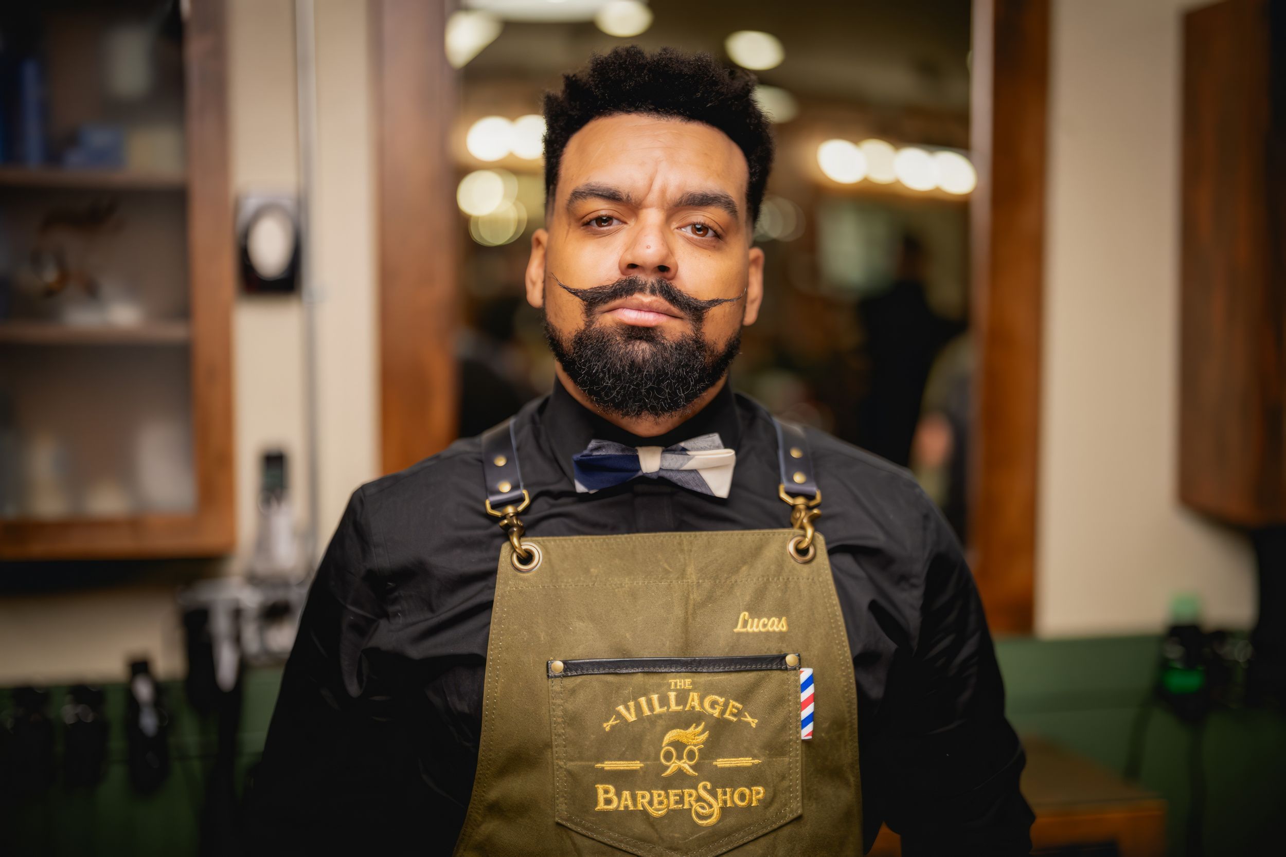 Barista wearing a brown apron and bow tie smiles warmly at the camera in a coffee shop setting.