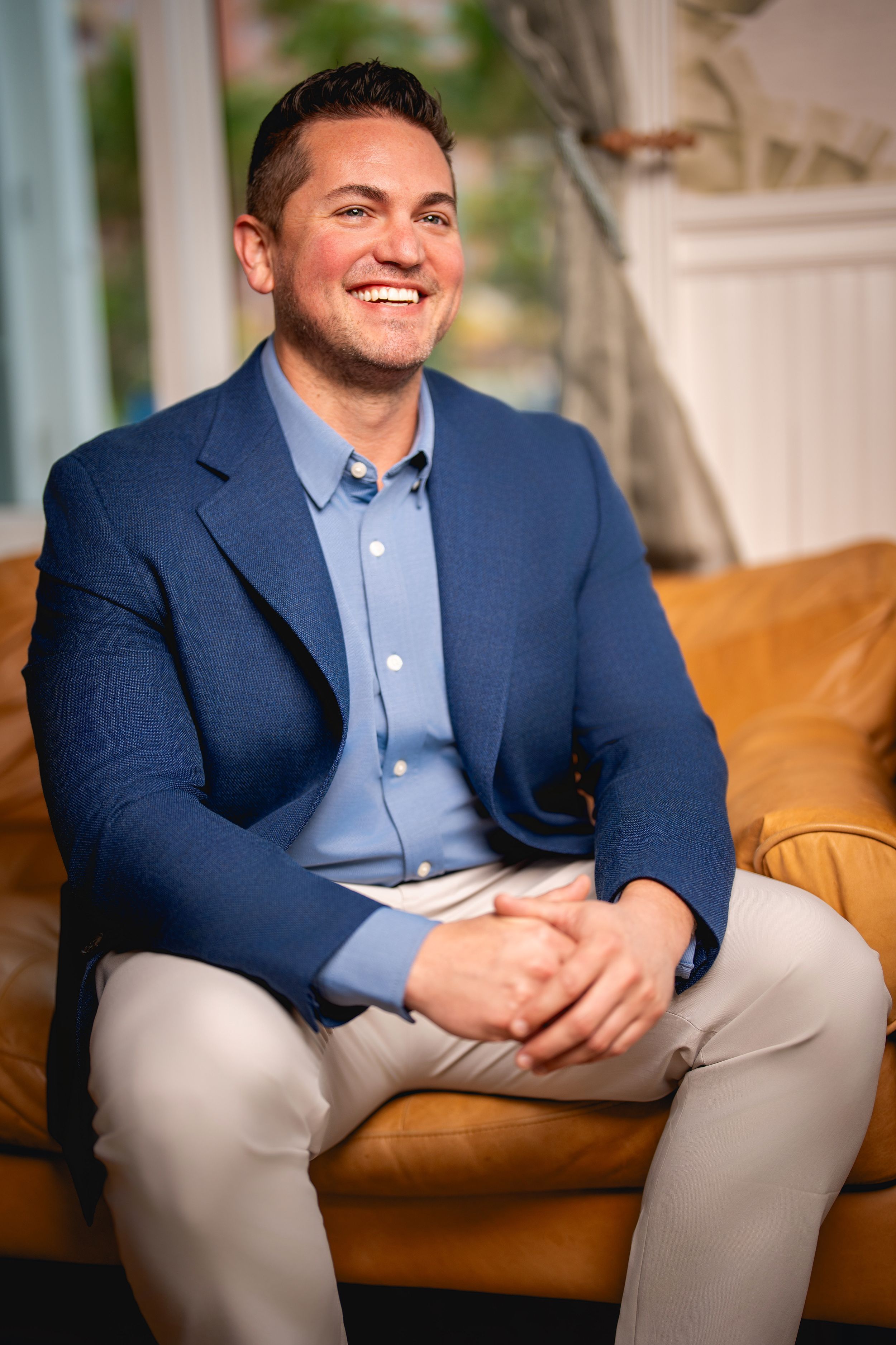 Professional portrait of someone wearing a navy blazer and light blue shirt sitting on a brown leather couch and smiling.