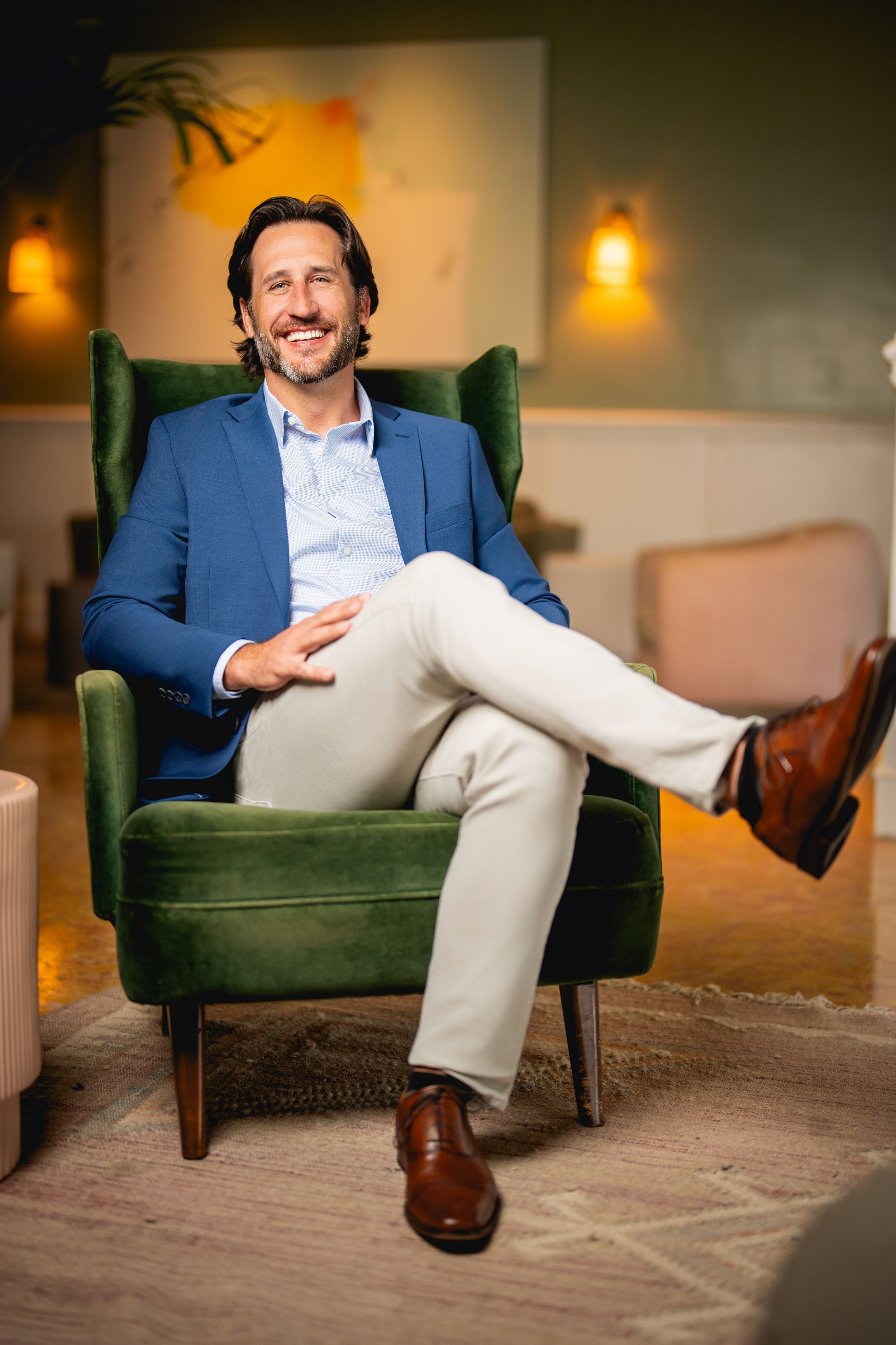 A smiling person sits relaxed in a green velvet armchair wearing a blue blazer, white shirt and tan pants.