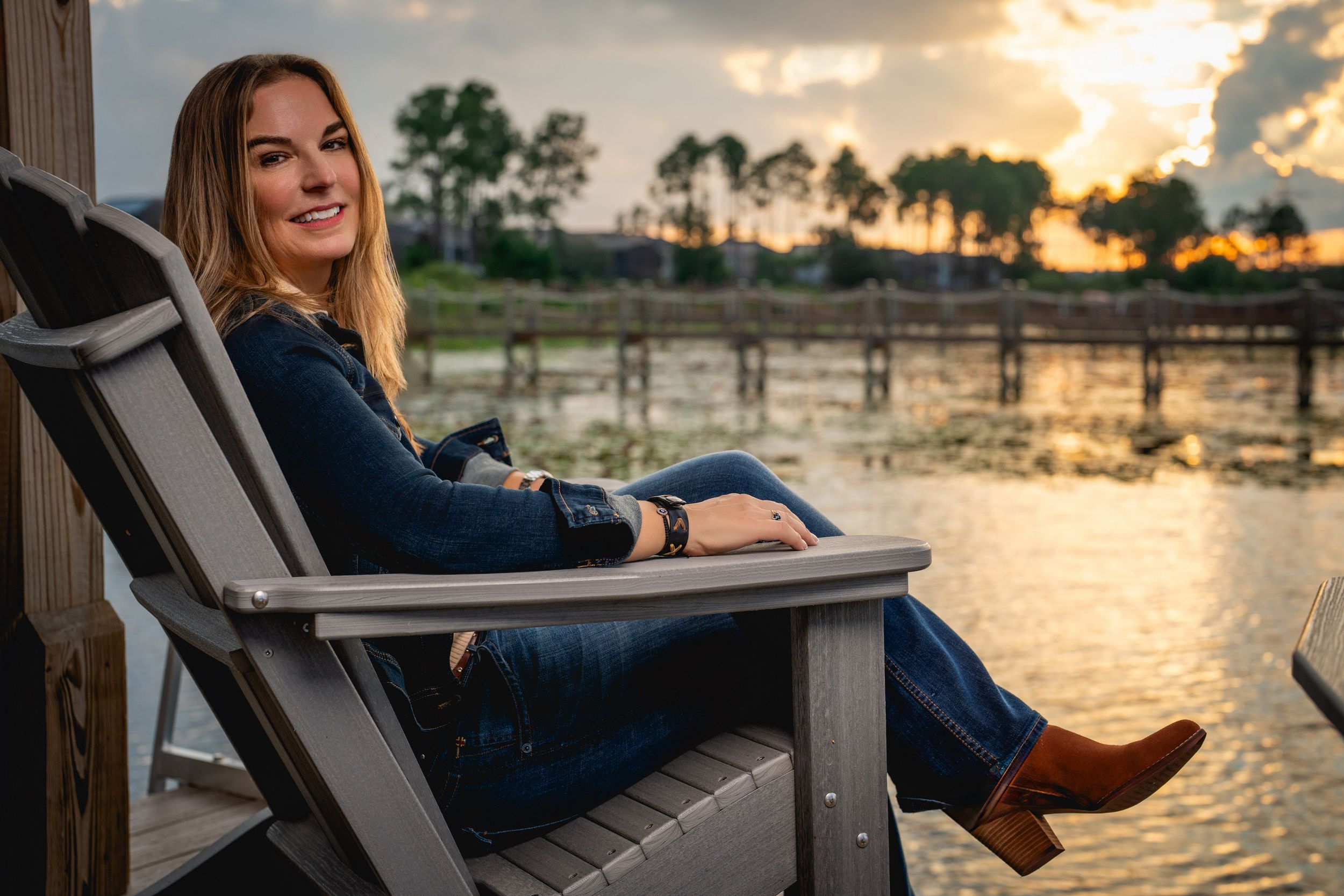 A person relaxes in an Adirondack chair by a tranquil lake during golden hour sunset.