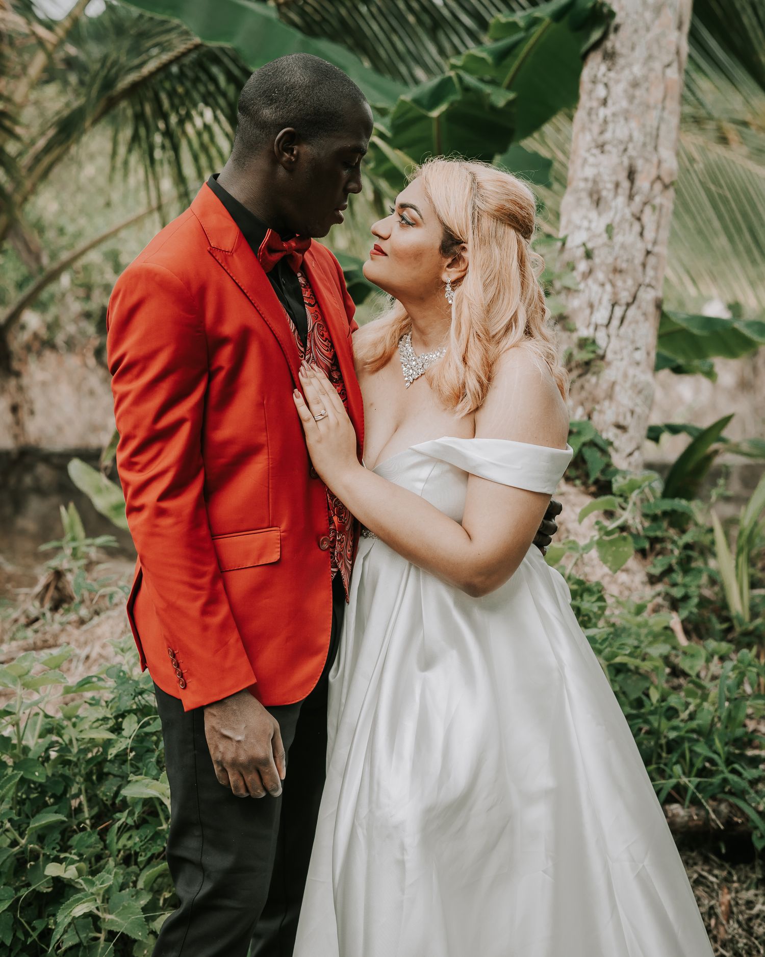 Romantic couple in tropical garden with groom in red jacket and bride in white gown.