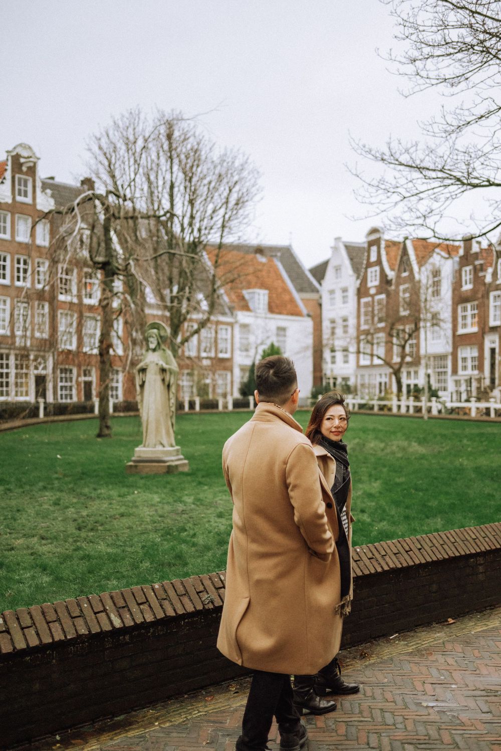 Two people in winter coats stand near statue in historic Amsterdam courtyard surrounded by Dutch architecture.