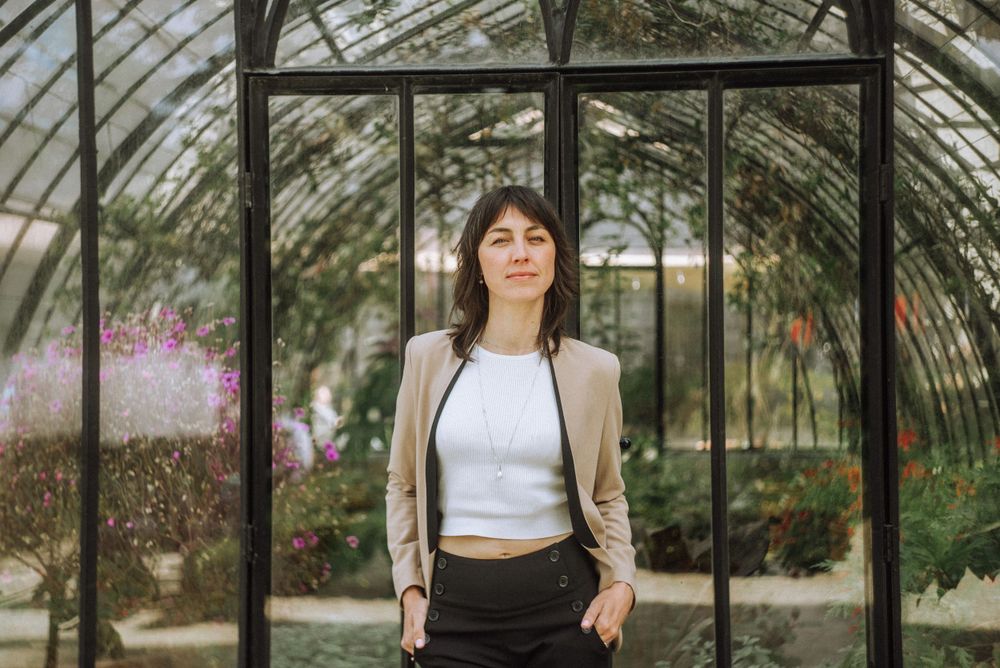 Person in a white crop top and beige blazer poses inside a greenhouse with natural lighting.