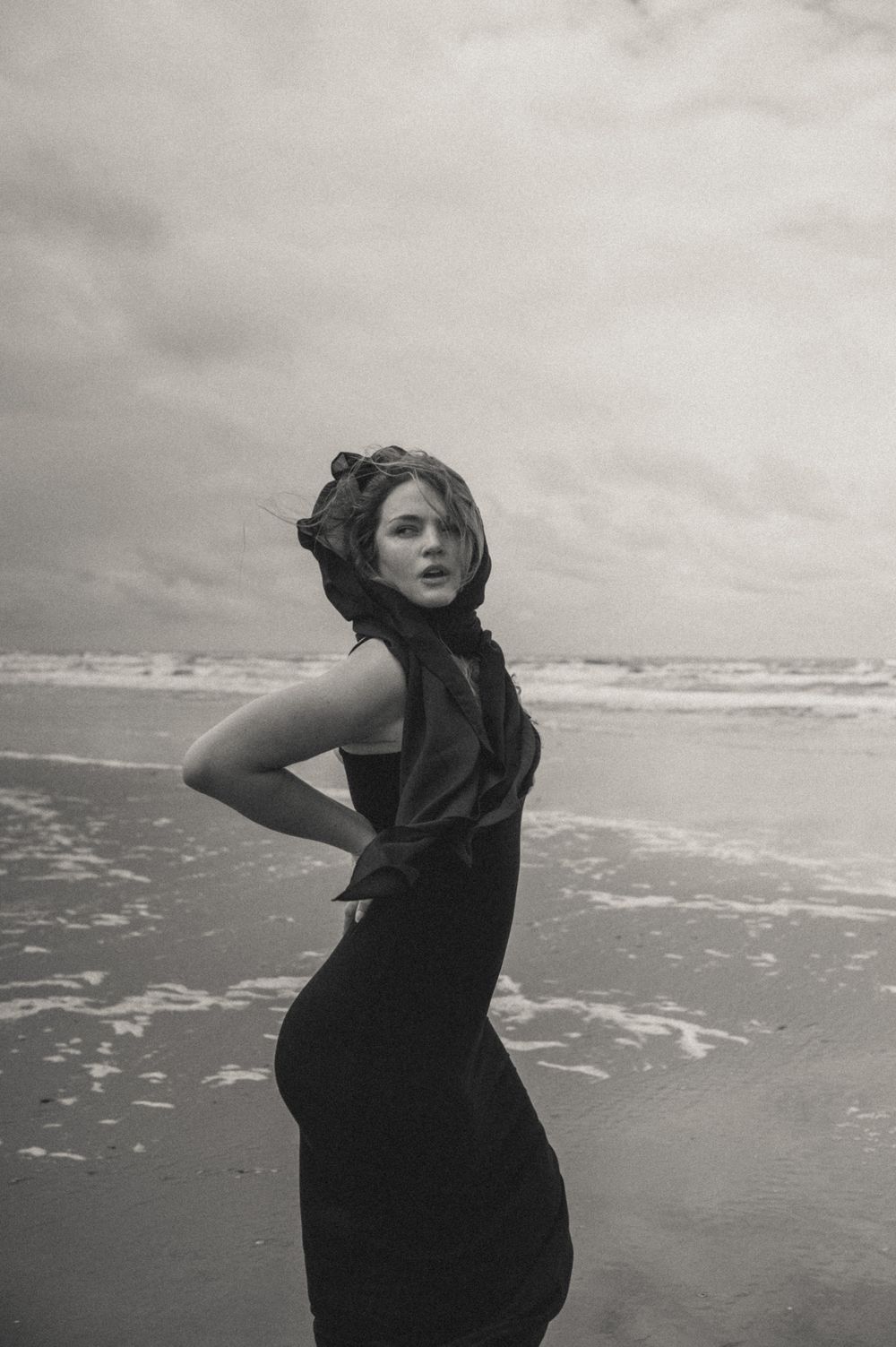 Dramatic black and white portrait captured against ocean waves and cloudy sky.