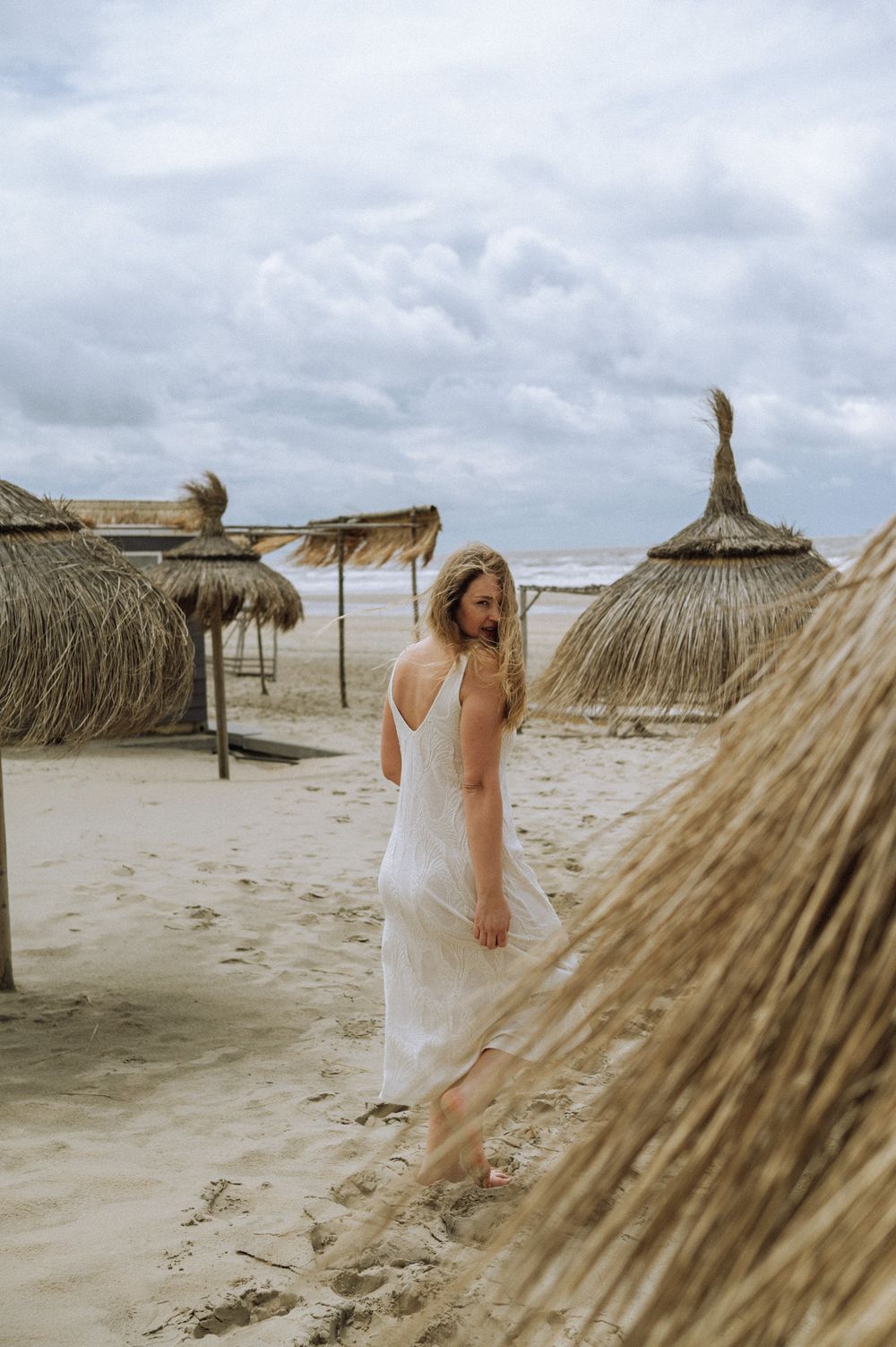 Beach scene with thatched umbrellas and white dress against cloudy coastal backdrop.