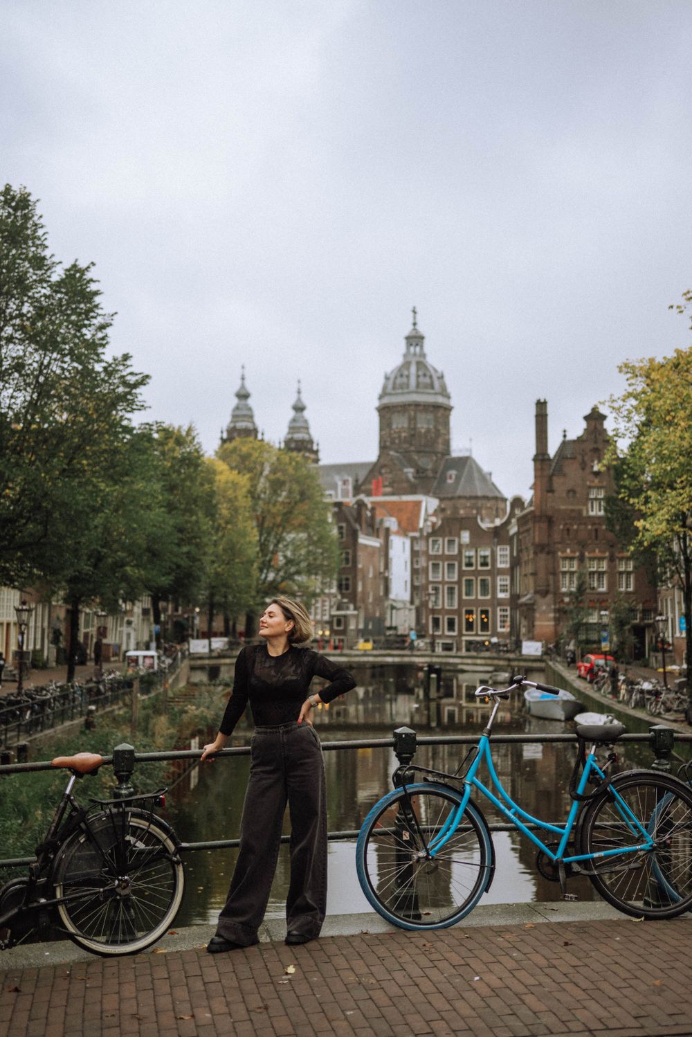 Person stands on Amsterdam bridge with blue bicycle and historic church domes in background.