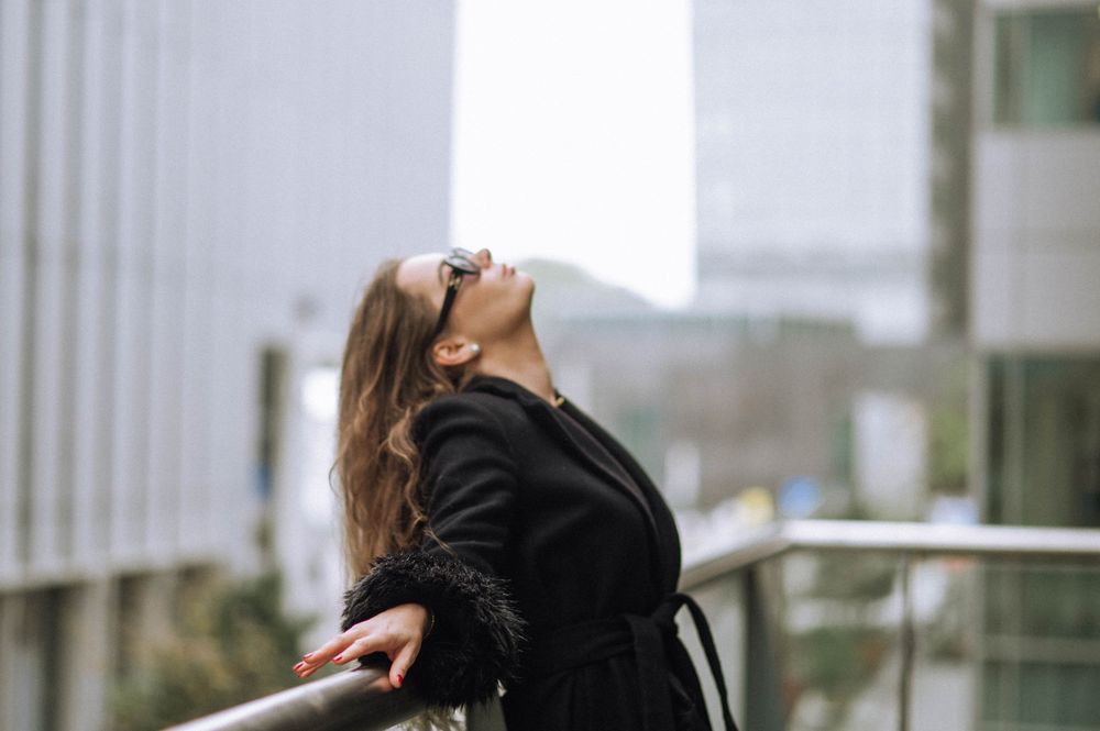 Person in black coat tilts head back while standing on modern balcony.