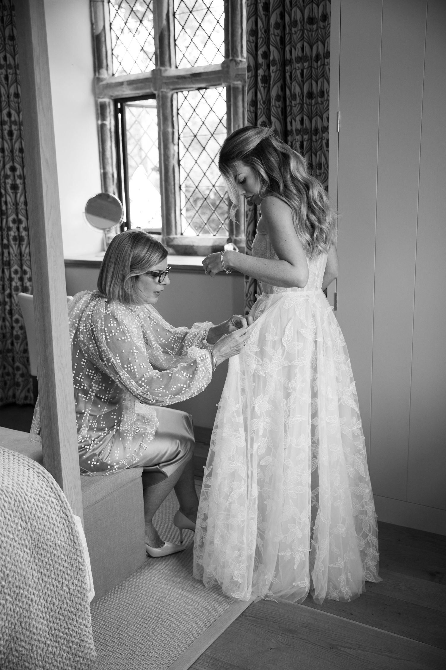 A bride in a lace wedding dress getting help with final preparations in a room with vintage windows.