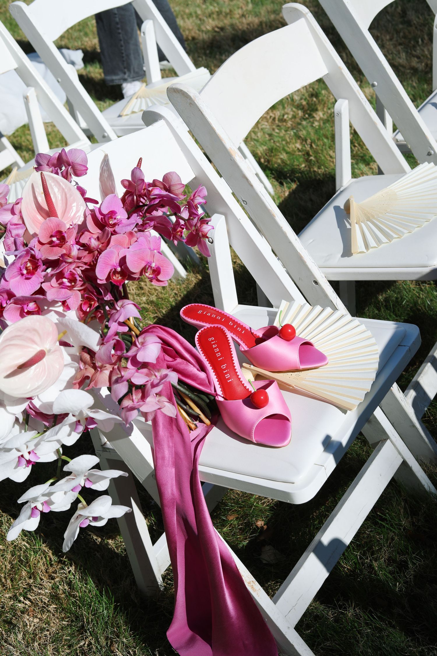 Pink orchids and matching high heel shoes displayed on white folding chairs outdoors.