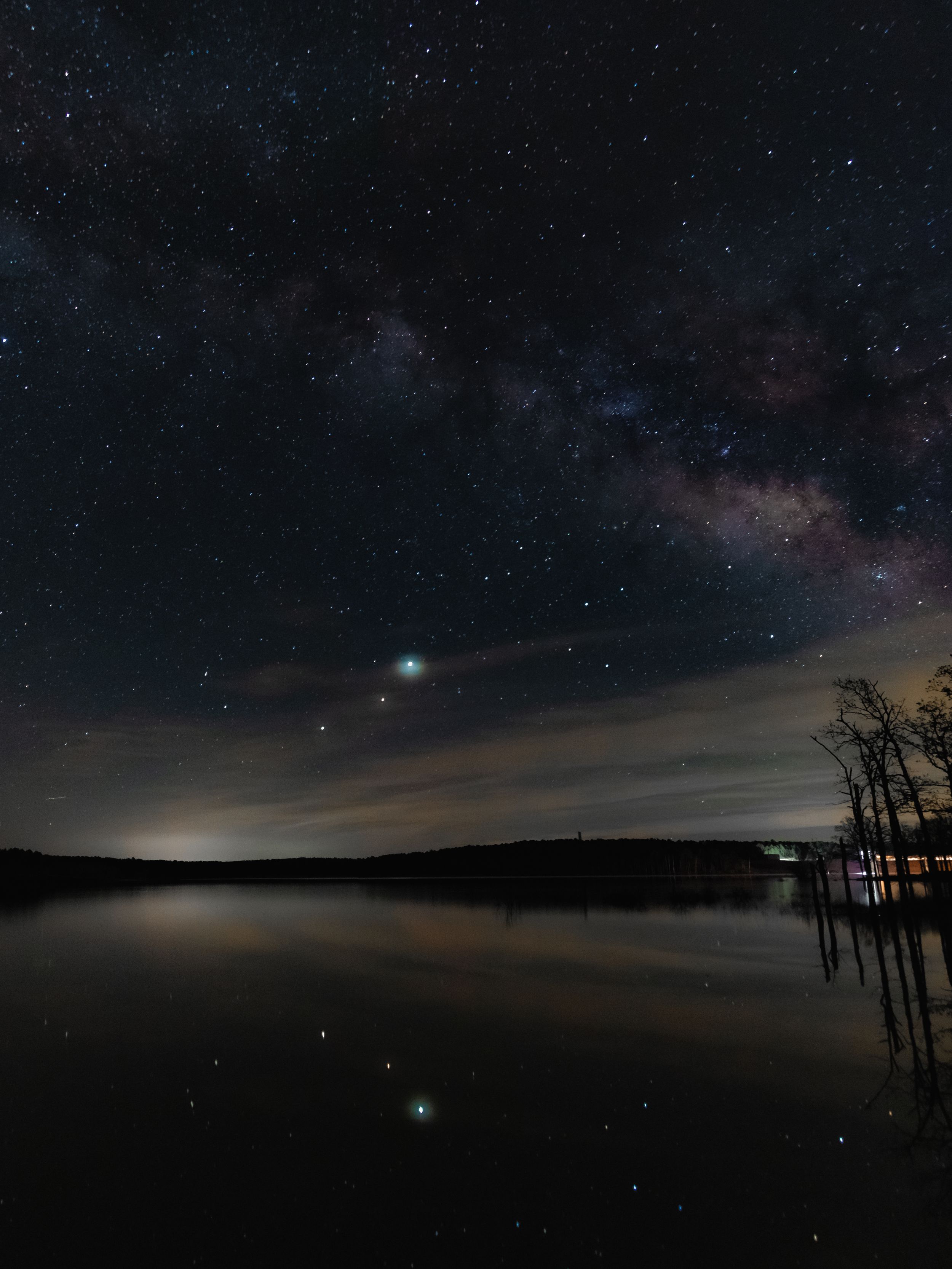 Milky Way galaxy reflects in still lake waters under a starry night sky with silhouetted trees.