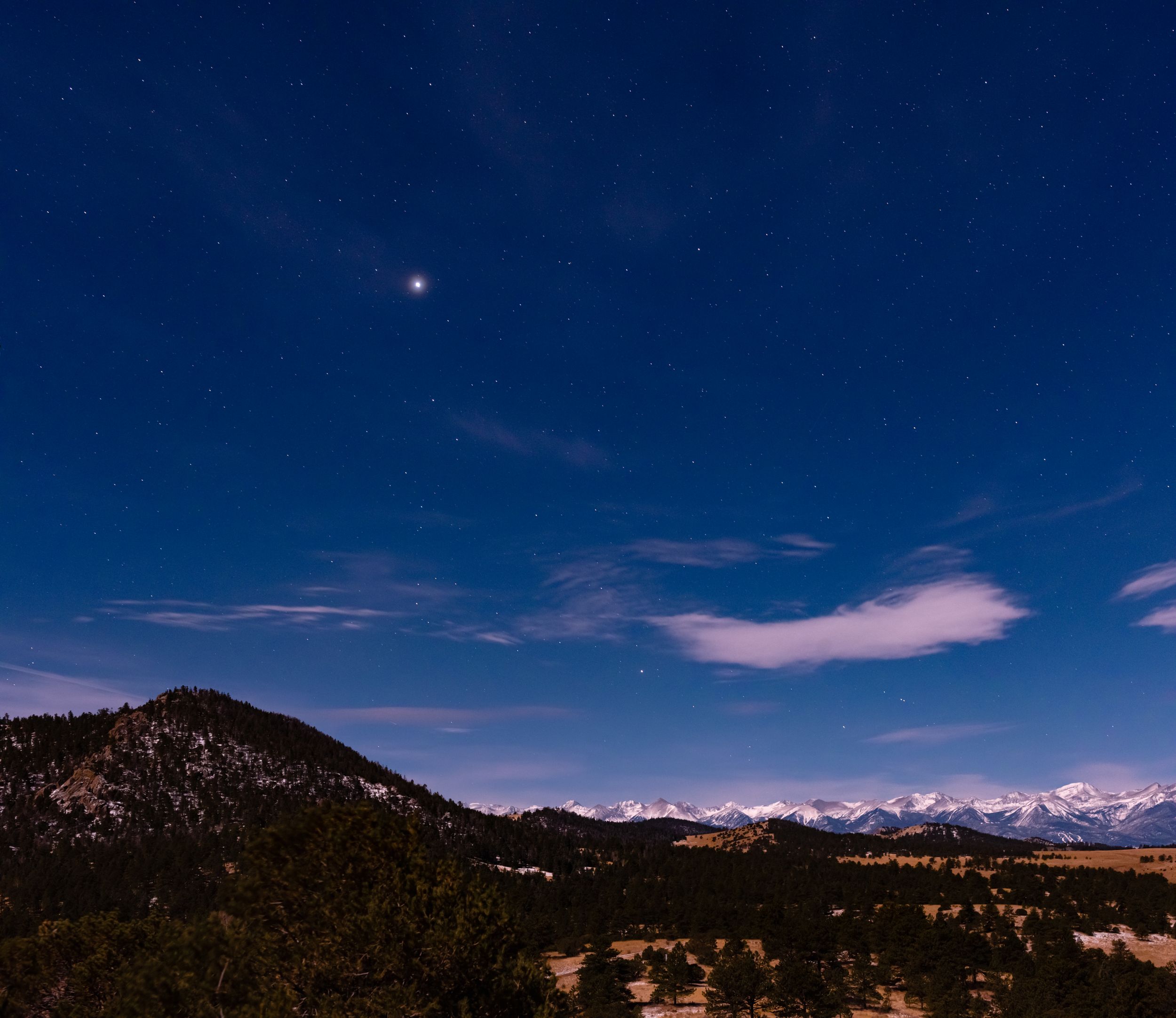 Night sky over mountain range with bright celestial object and wispy clouds.