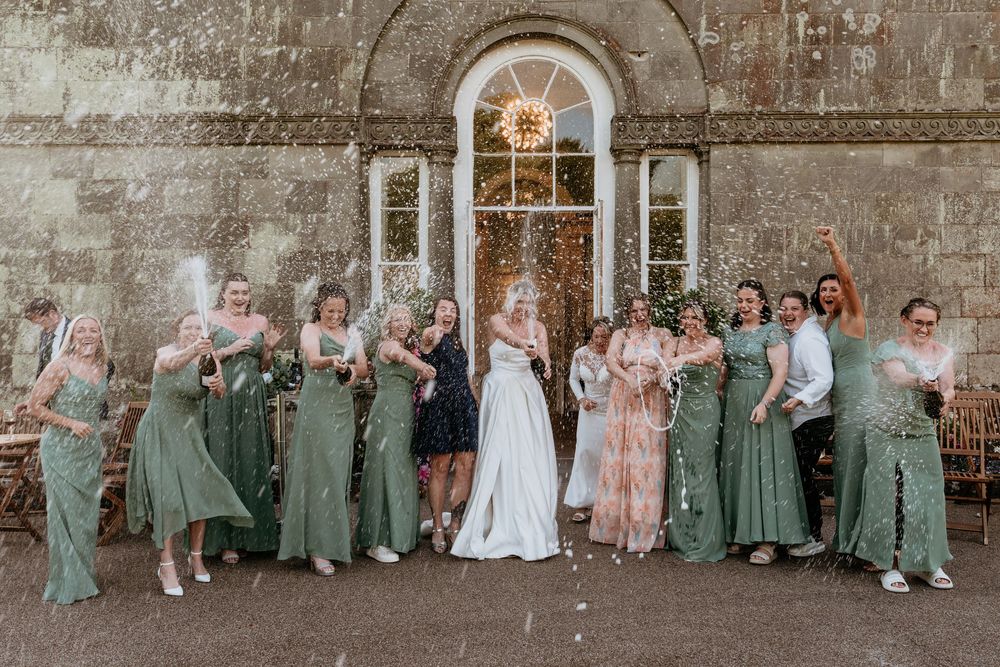 Wedding party in coordinated green and pink dresses poses in front of a stone wall for group photos.