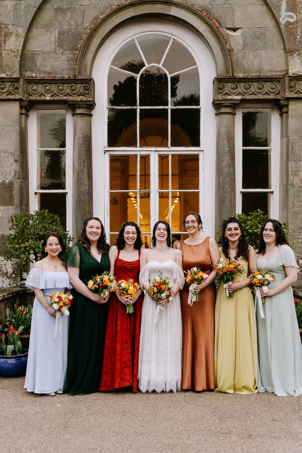 A wedding party in colorful mix-and-match dresses stands in front of a stone building with arched windows and columns.