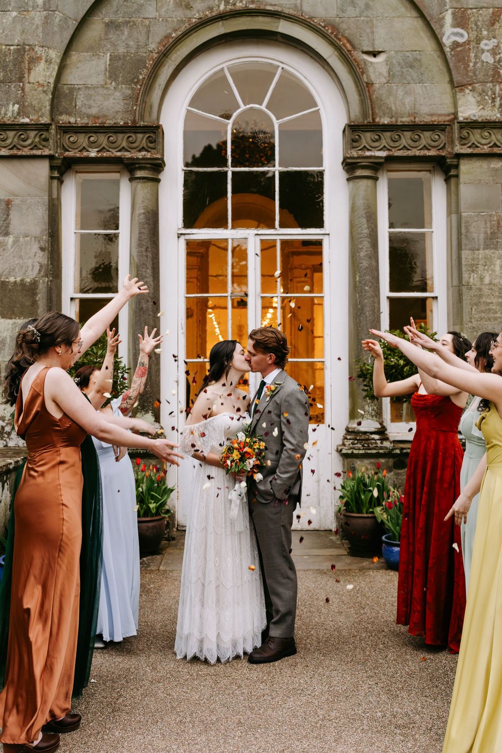 Wedding party members in colorful dresses celebrate joyfully outside a stone building with elegant arched windows.