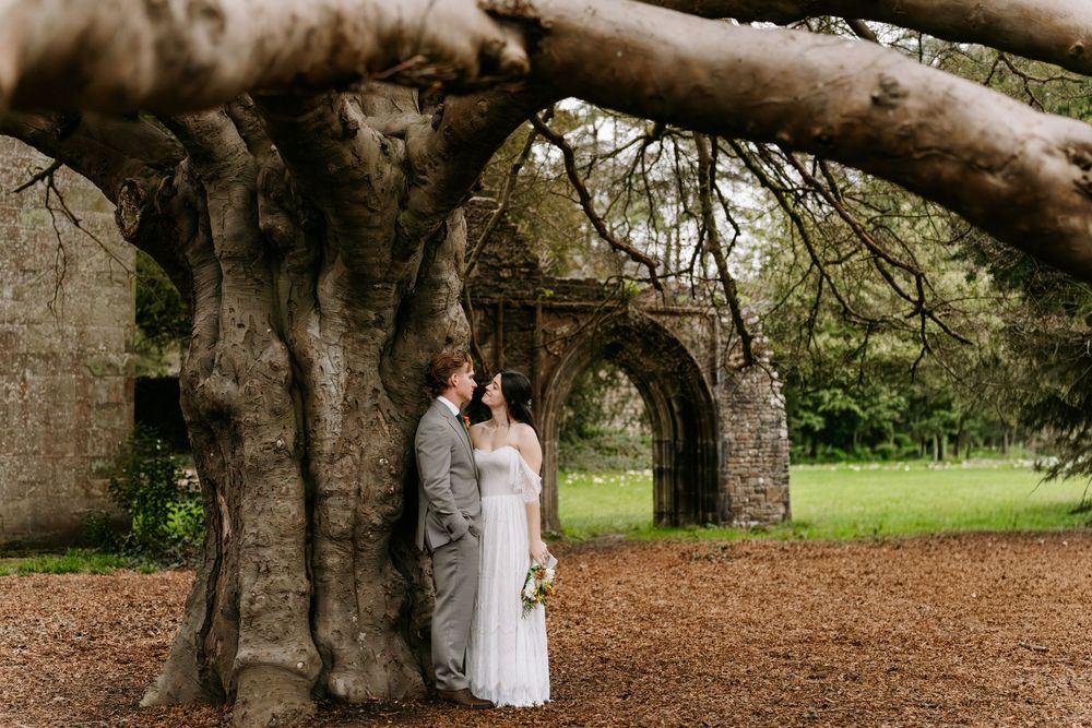 A couple stands beneath a massive tree trunk and stone archway in a romantic garden setting.