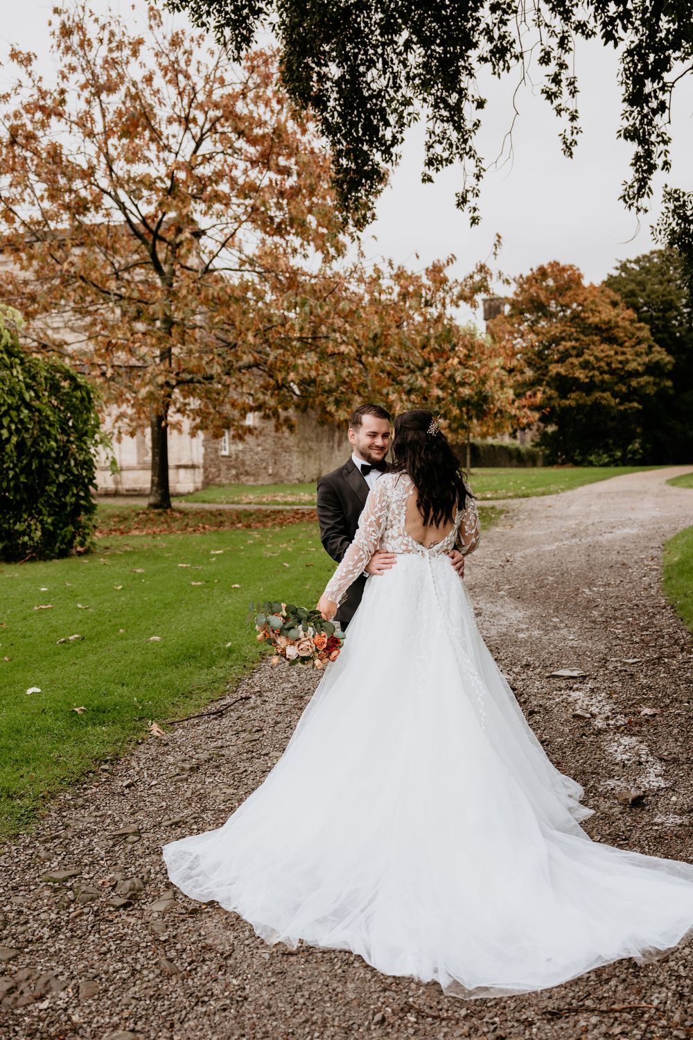 A couple in wedding attire walks along a gravel path surrounded by autumn trees with orange foliage.