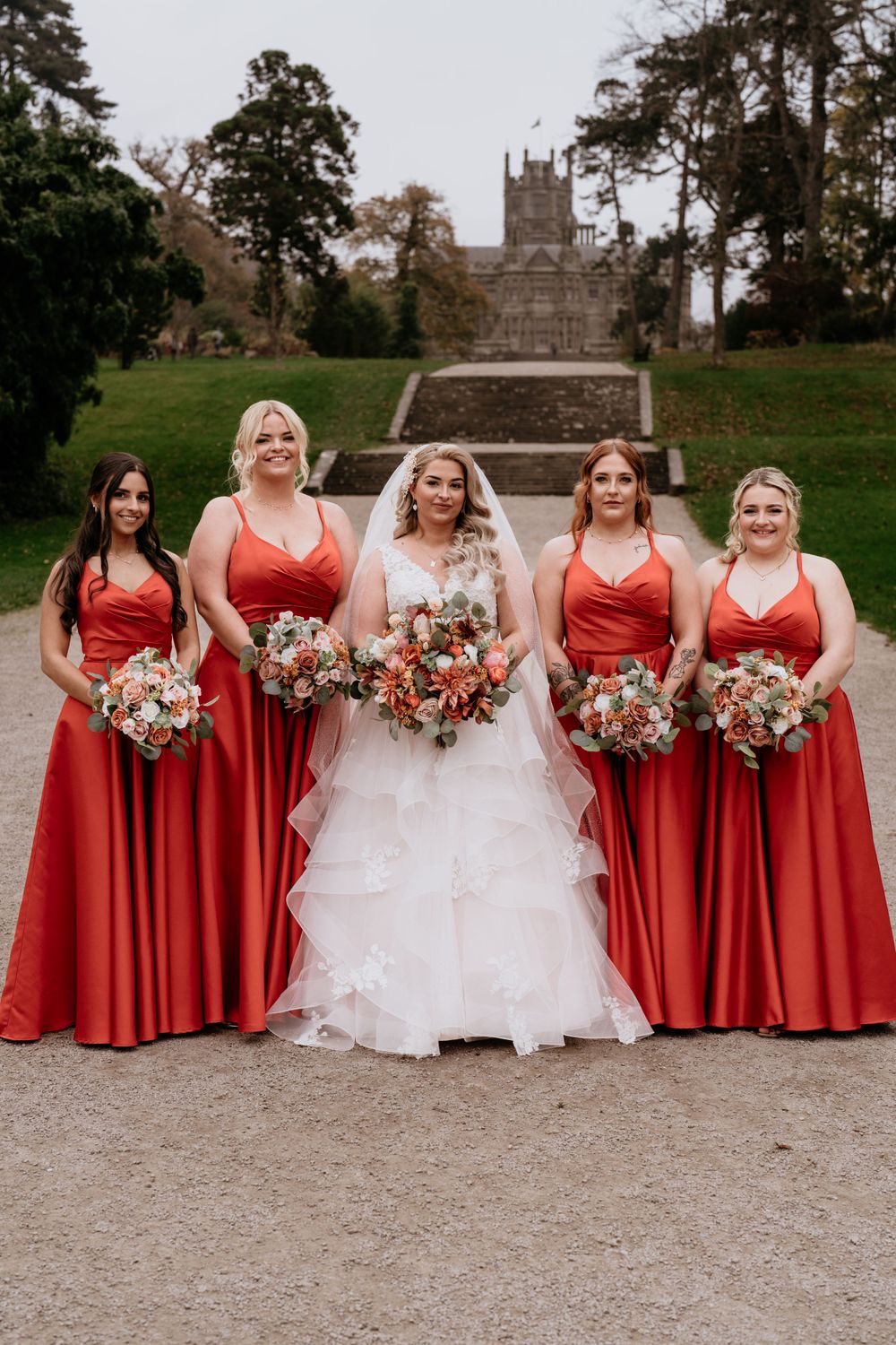 A wedding party with bridesmaids in red dresses holding bouquets pose in front of a historic castle on a gray day  at The Orangery, Margam