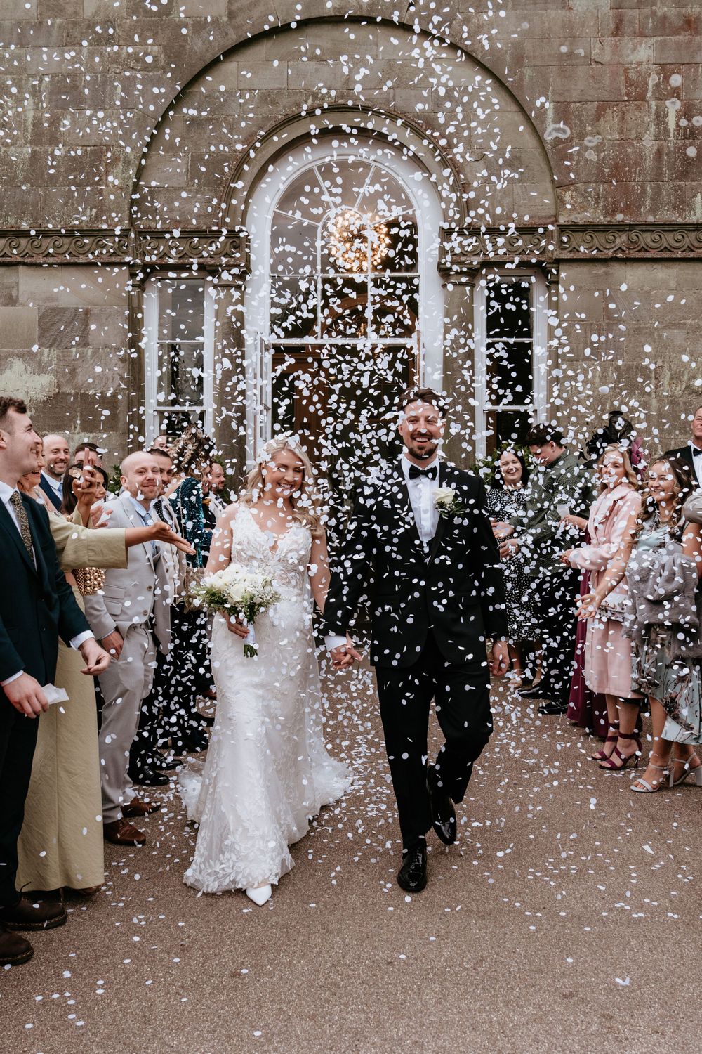 Wedding couple exits church as guests shower them with white confetti against brick building backdrop at the Orangery Margam